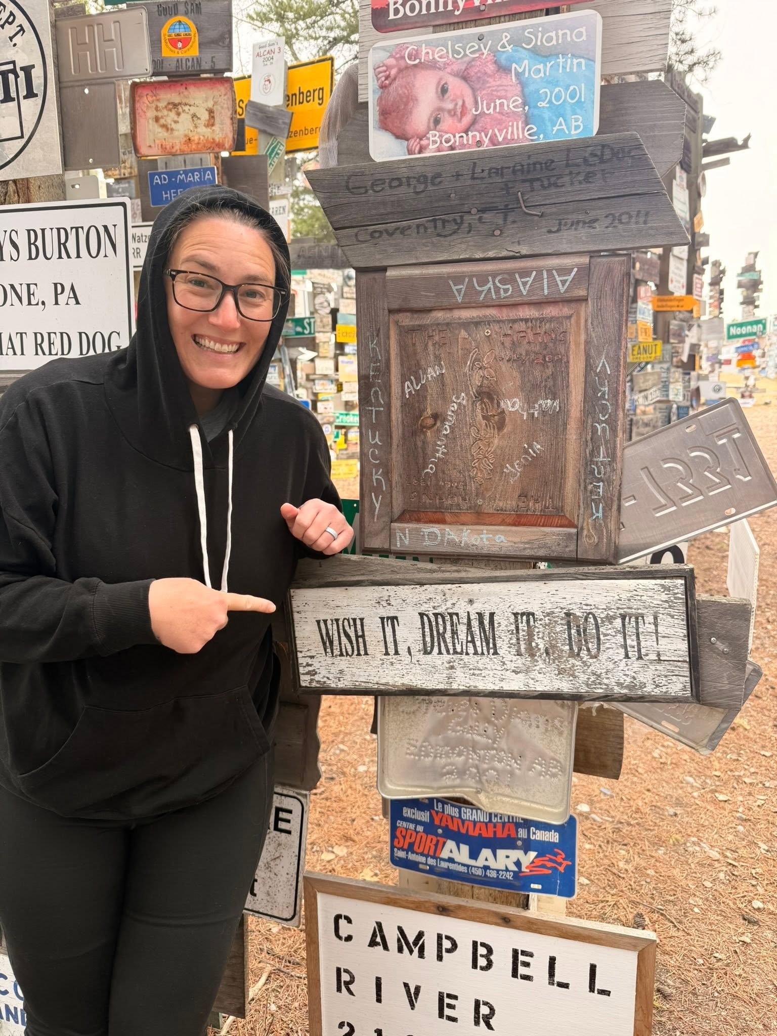 Jennifer beside a Wish it, Dream it, Do it sign in the Watson Lake Sign Post Forest in Yukon
